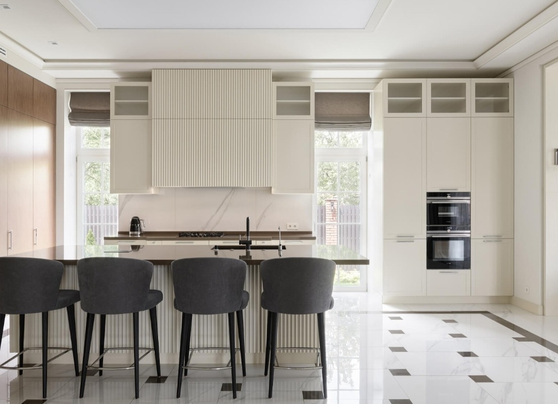 Nowoczesna kuchnia z wyspą i czterema szarymi hokerami, białe szafki z ryflowanym frontem nad blatem, marmurowy backsplash, podłoga w biało-czarną szachownicę.