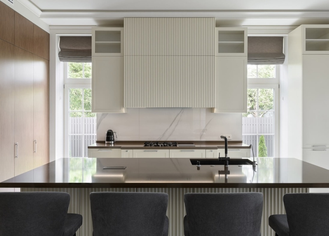 Nowoczesna kuchnia z wyspą, kremowe szafki z frezowanymi frontami, marmurowy backsplash, czarne akcenty (bateria, czajnik), szare krzesła barowe i duże okna z roletami rzymskimi.