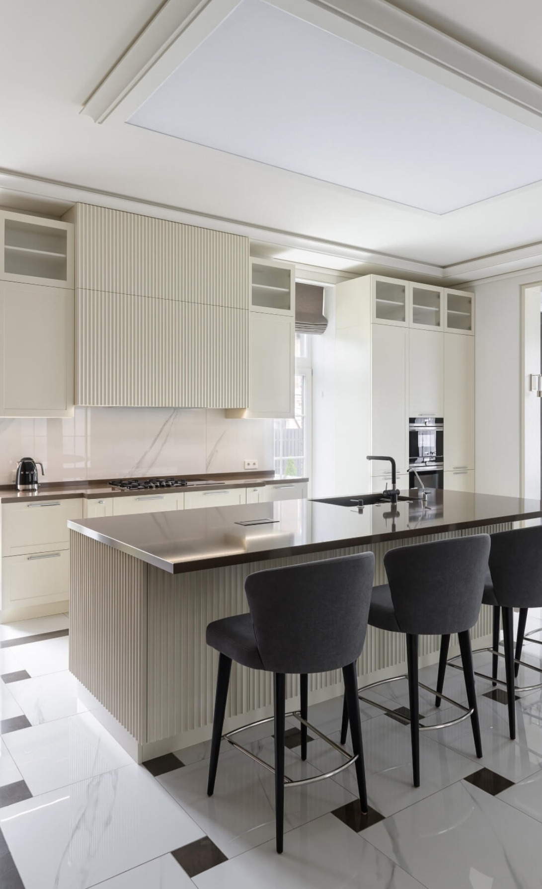 Elegancka kuchnia w jasnych barwach z wyspą i trzema szarymi hokerami, marmurowa podłoga z ciemnymi akcentami, nowoczesne oświetlenie sufitowe.