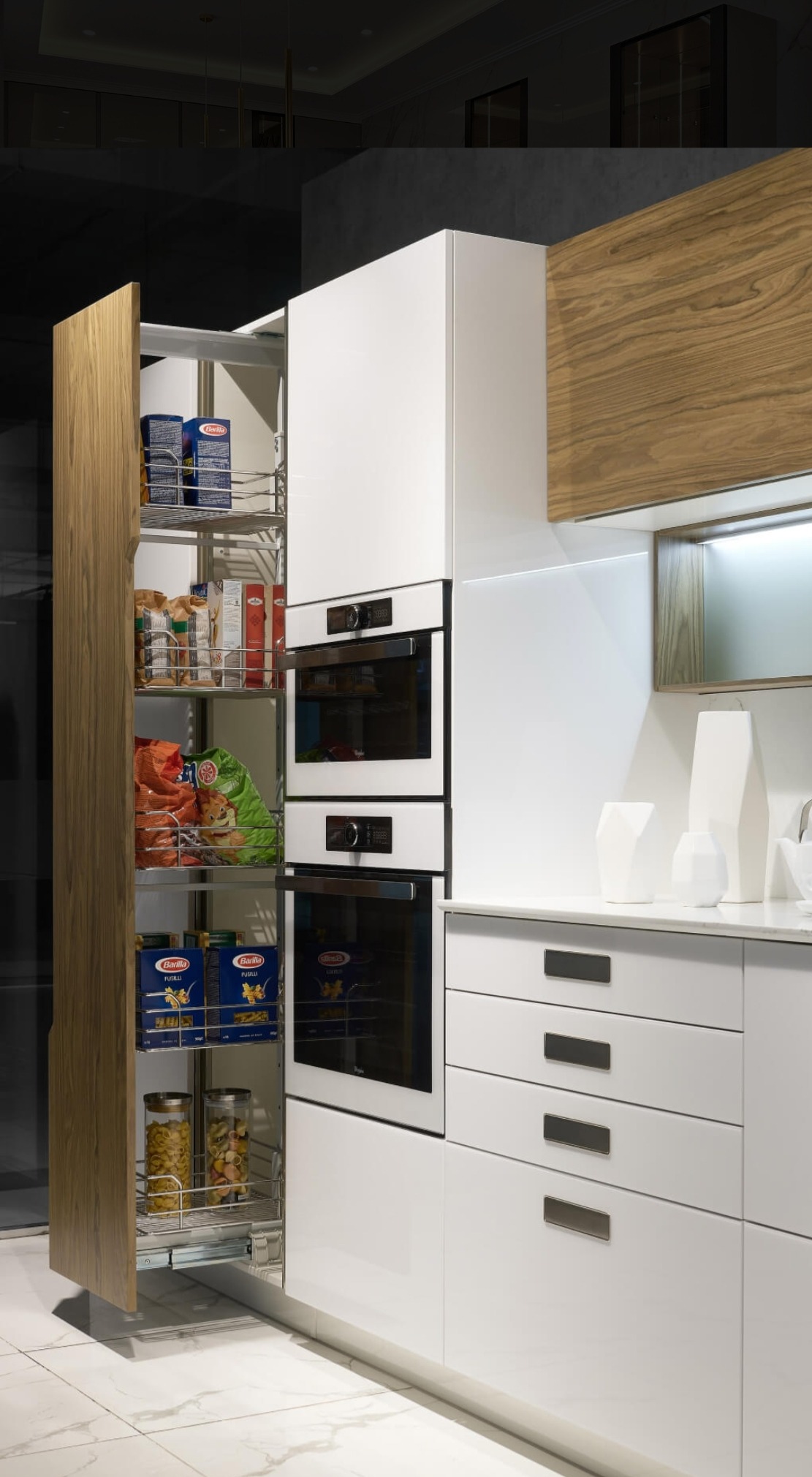 Nowoczesna biała kuchnia z wysuwaną szafką cargo wypełnioną produktami spożywczymi, obok zabudowane piekarniki i białe szafki z szufladami.