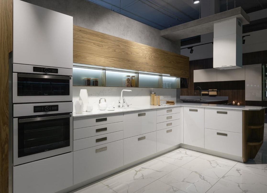 Nowoczesna, biała kuchnia z drewnianymi akcentami, podświetlanymi szafkami górnymi i marmurową podłogą, piekarnik w zabudowie.