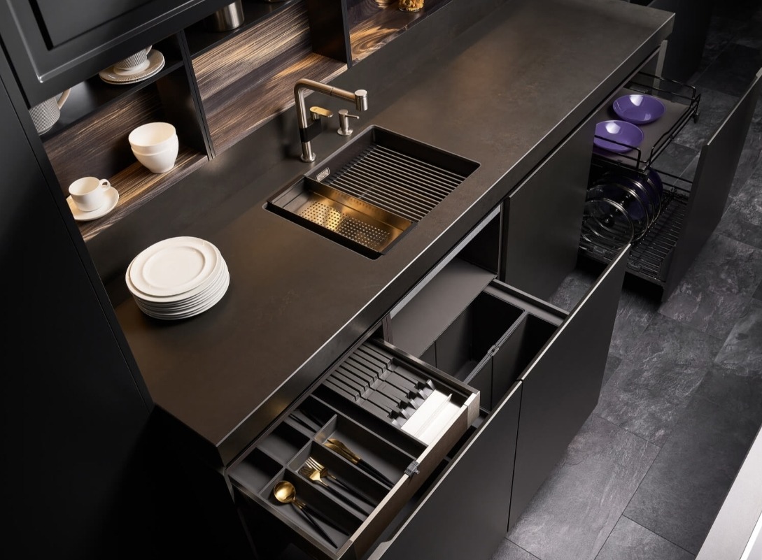 Nowoczesna, ciemna zabudowa kuchenna w Warszawie z otwartymi szufladami na sztućce i naczynia, zintegrowany zlew z ociekaczem i minimalistyczna armatura.