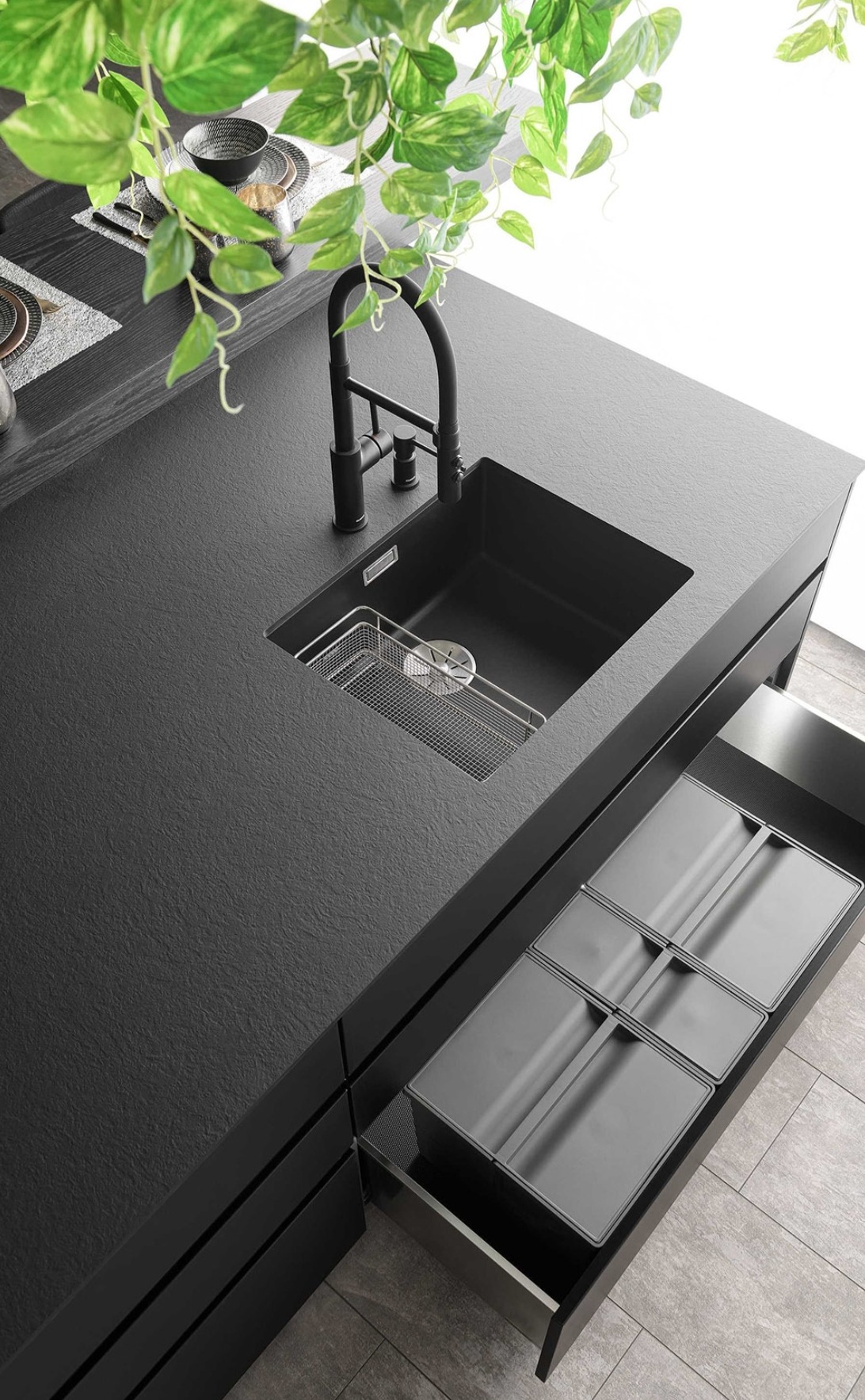 Czarny, minimalistyczny blat kuchenny z wbudowanym czarnym zlewem, metalowym ociekaczem i baterią, otwartą szufladą z organizerami na sztućce, oraz zieloną rośliną w tle.