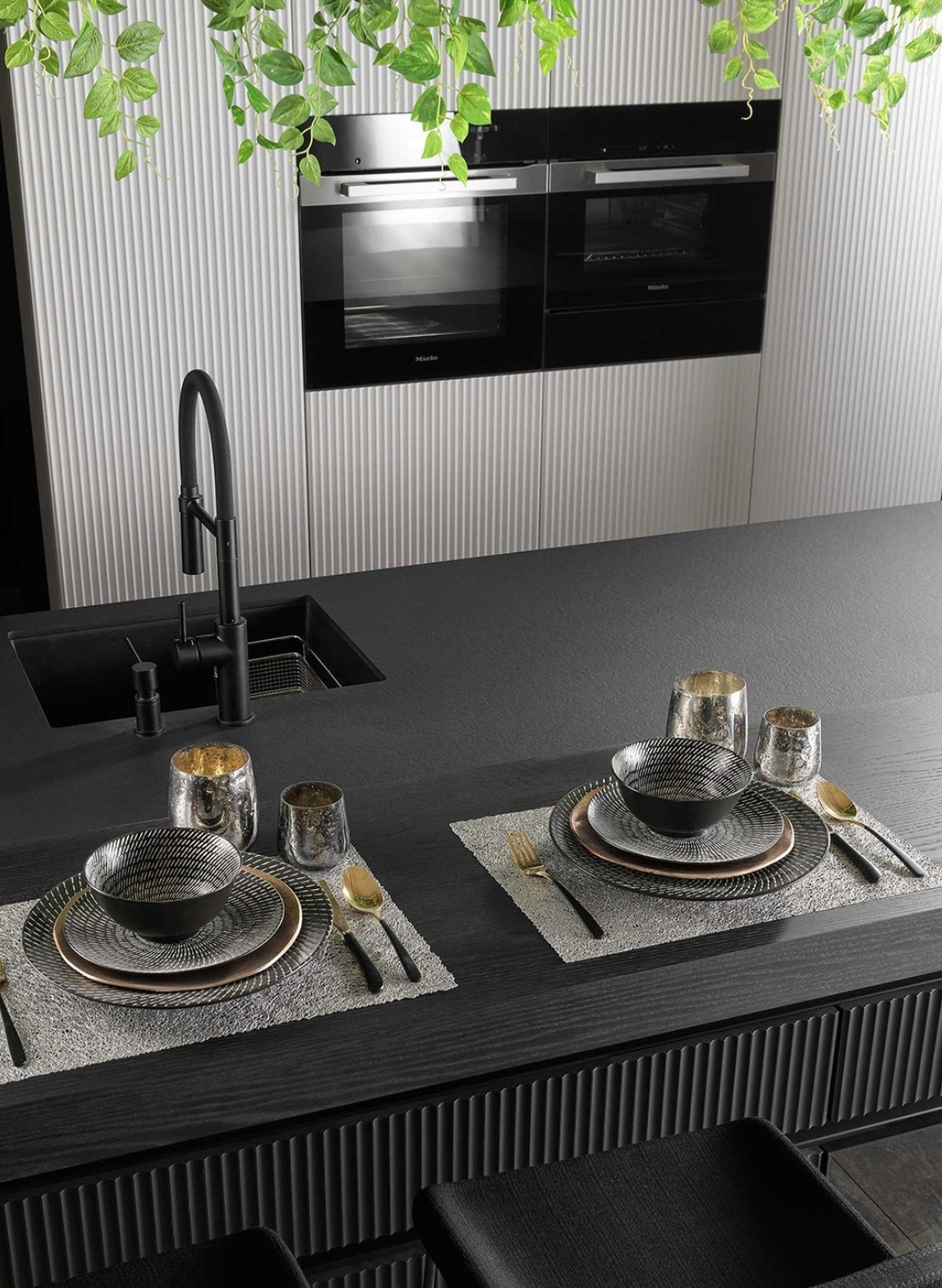 Elegancka, czarna kuchnia z wbudowanymi piekarnikami Miele, czarnym zlewem i baterią, oraz nakrytym stołem dla dwóch osób z dekoracyjnymi naczyniami i złotymi sztućcami, dopełniona zwisającymi...
