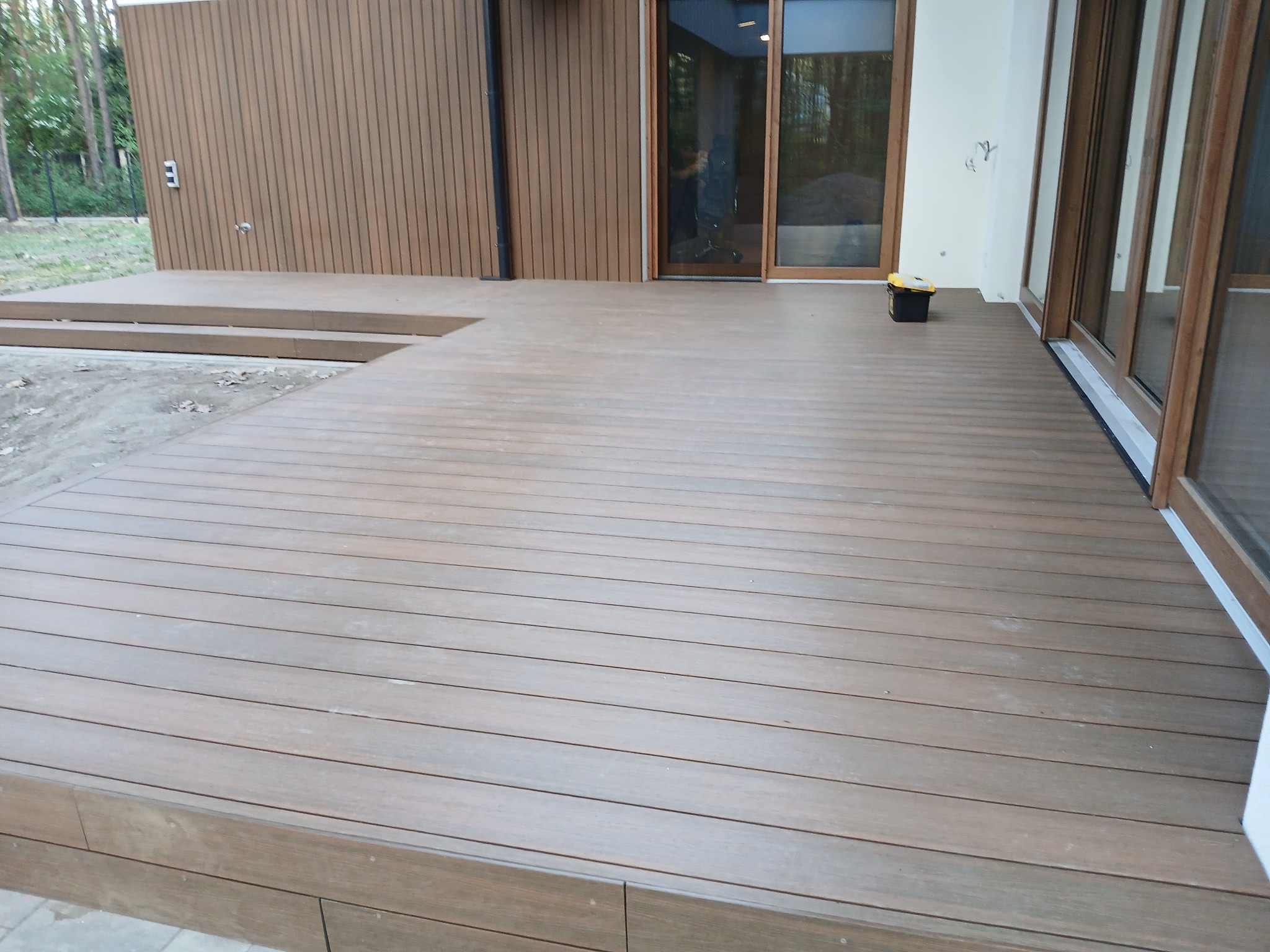 Kompozytowy taras w odcieniach brązu, z widocznymi deskami i subtelnymi detalami wykończenia, przylegający do nowoczesnego budynku z dużymi oknami.