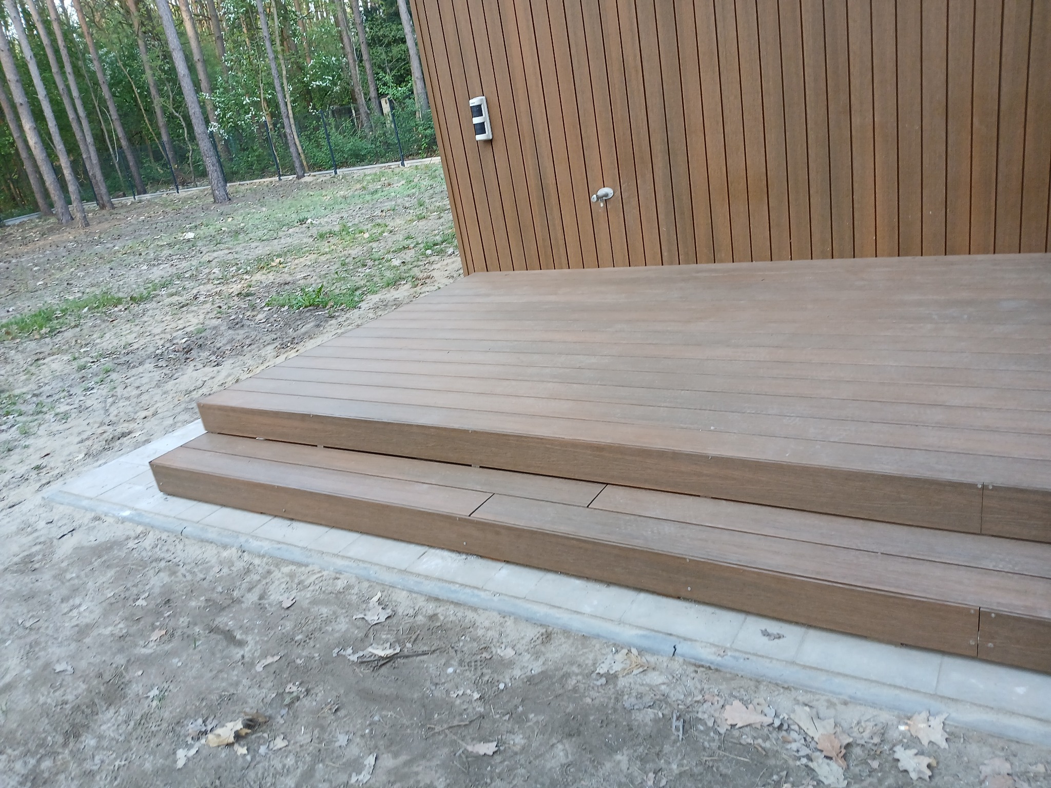 Nowoczesny taras kompozytowy z dwustopniowymi schodami, wykończony płytami chodnikowymi, na tle lasu. Eleganckie detale i naturalne otoczenie.