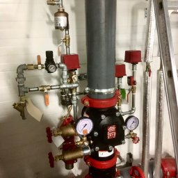 INSTALACJE WOD.- KAN. GAZ i C.O. Kamil Kruk - Złożony system rur i zaworów z manometrami w kolorach szarym, czarnym i czerwonym, z widoczną drabinką aluminiową po prawej stronie, prawdopodobnie element instalacji przeciwpożarowej.