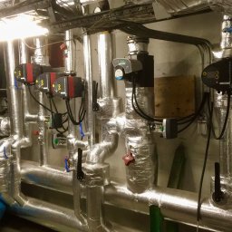 INSTALACJE WOD.- KAN. GAZ i C.O. Kamil Kruk - Izolowane rury grzewcze z pompami obiegowymi Grundfos i zaworami, widoczne elementy instalacji w piwnicy, w tle czerwony zbiornik wyrównawczy, instalacja po montażu.