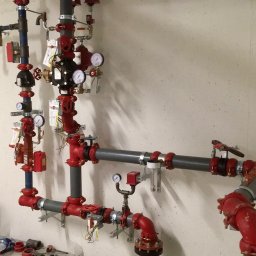 INSTALACJE WOD.- KAN. GAZ i C.O. Kamil Kruk - Kompleksowy system rur, zaworów i manometrów, pomalowanych na czerwono i szaro, zamontowany na ścianie w Skokach. Widoczne przewody elektryczne i elementy montażowe.