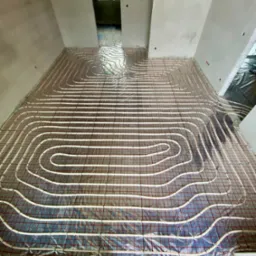 Rozłożona instalacja ogrzewania podłogowego na folii aluminiowej w surowym stanie, widoczne rury w kształcie meandrów i czerwona siatka mocująca, przygotowane do zalania wylewką.