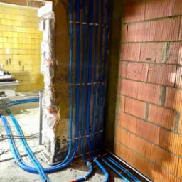 Niebieskie rury instalacji wodnej lub grzewczej, zamocowane do ściany z cegły i surowej ściany z resztkami tynku, widoczne pomarańczowe złączki i metalowe obejmy mocujące.