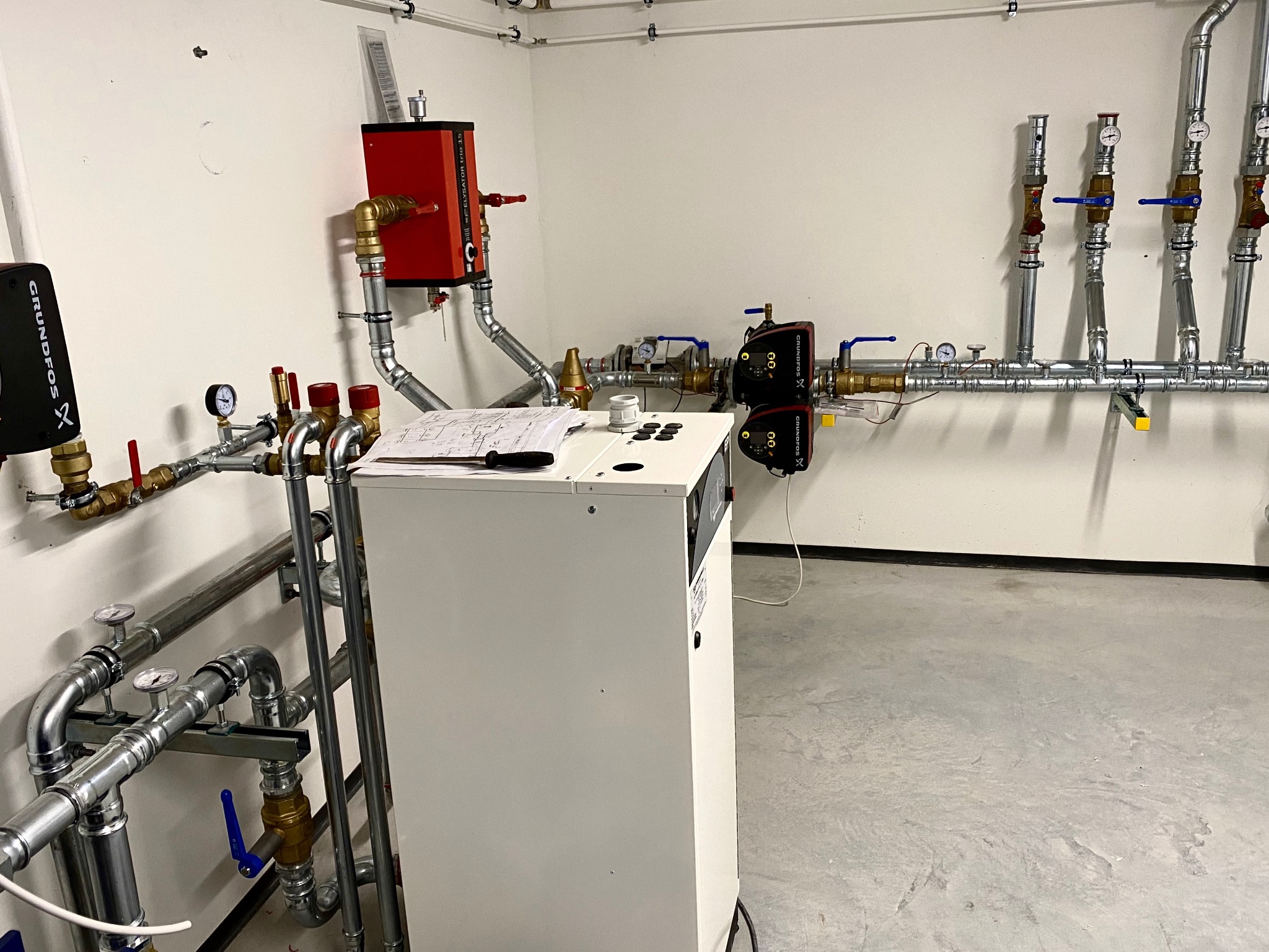 Węzeł cieplny: biała obudowa kotła z czarnymi przyciskami, obok instalacja z rur miedzianych i stalowych, zawory, manometry, pompy obiegowe Grundfos i schemat instalacji na obudowie.