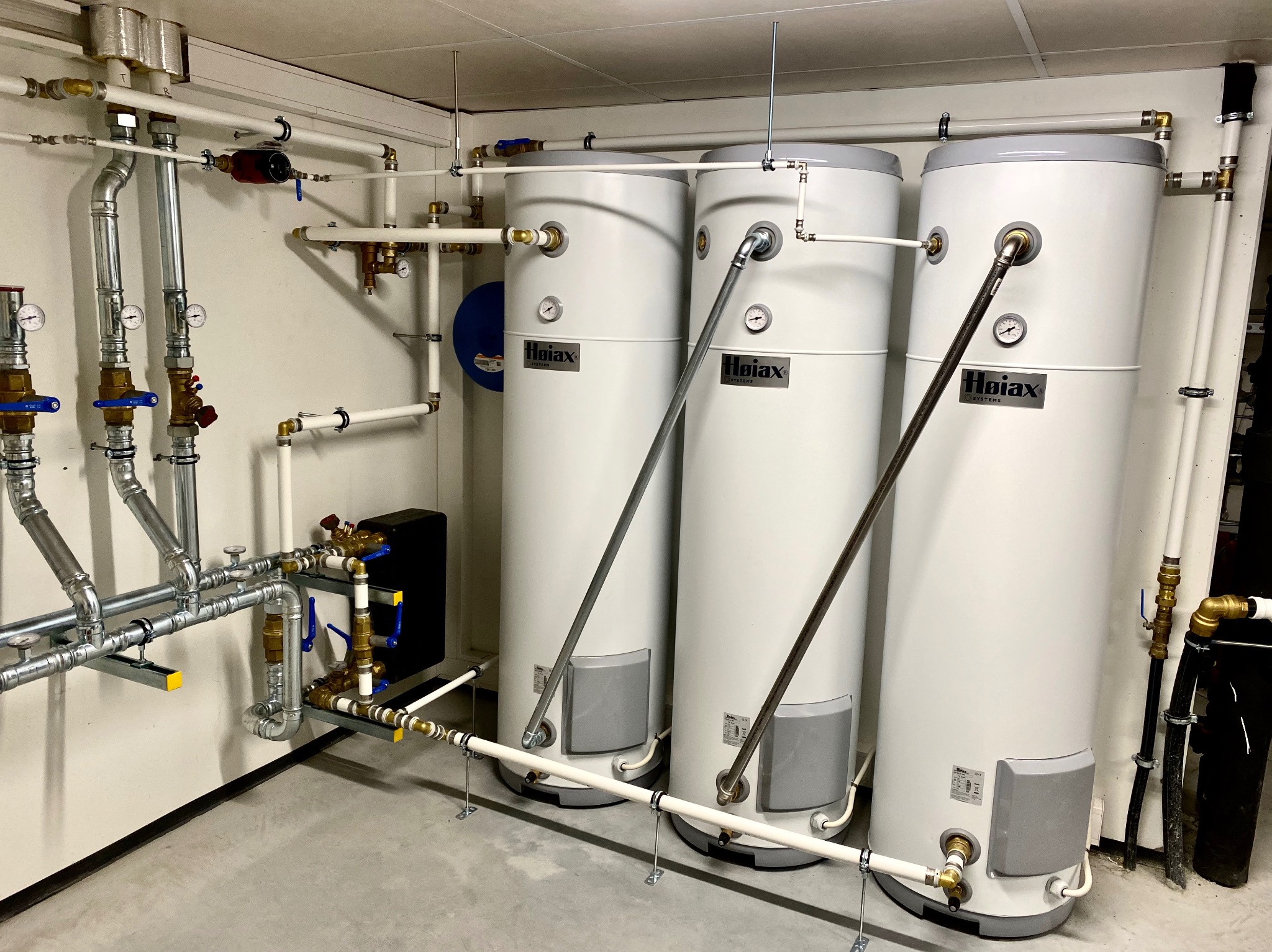 Trzy białe bojlery C.W.U. marki Hoiax, połączone rurami z zaworami, zamontowane w pomieszczeniu technicznym z szarą podłogą i białymi ścianami.
