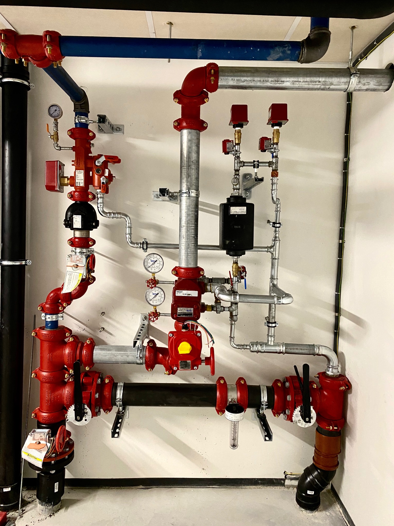 Złożona instalacja rur i zaworów w kolorach czerwonym, srebrnym i czarnym, zamontowana na białej ścianie, z widocznymi manometrami i elementami sterującymi.