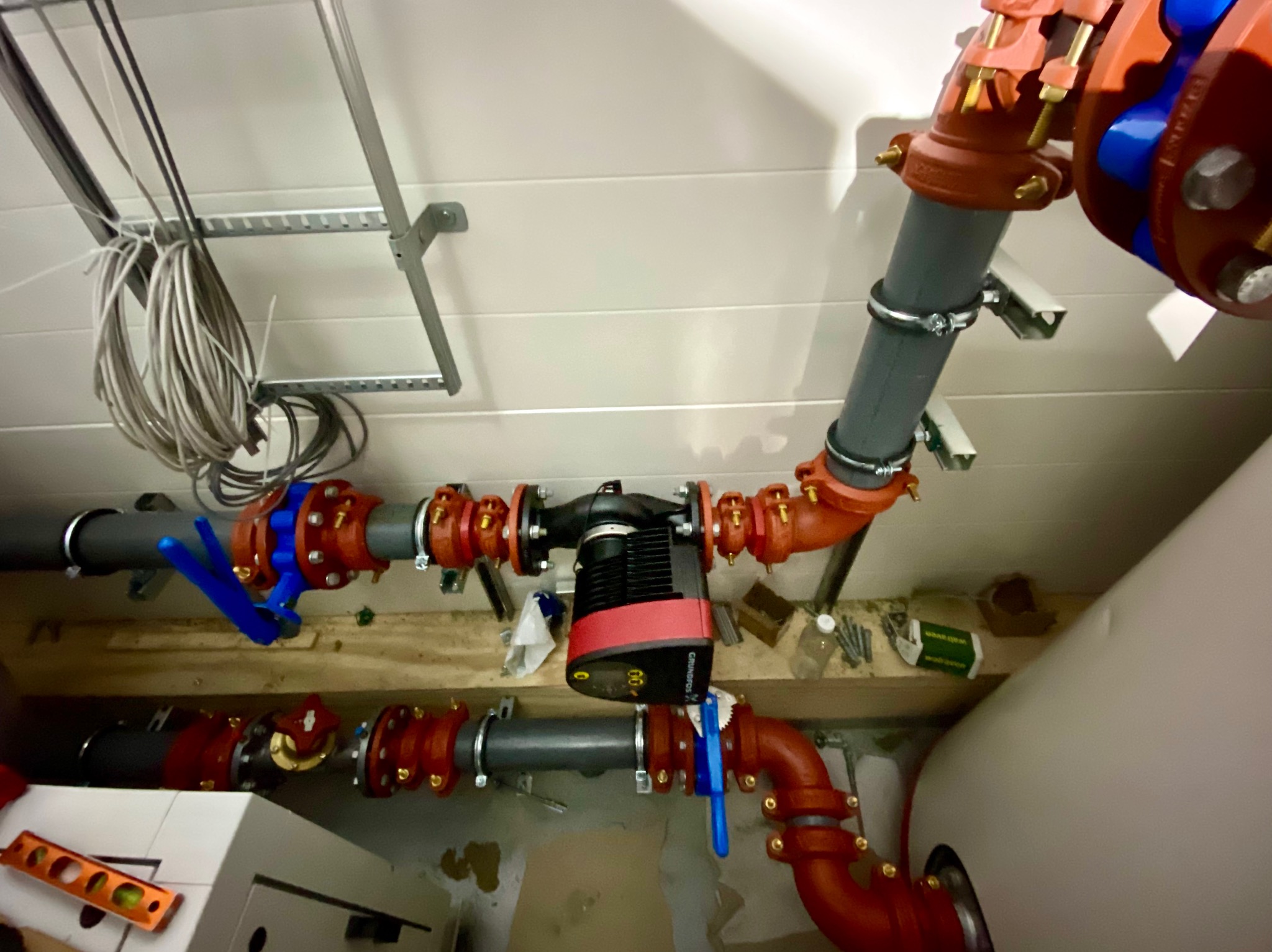 Instalacja hydrauliczna z pompą obiegową Grundfos i zaworami z niebieskimi uchwytami, rury w kolorze szarym i czerwonym, mocowane do ściany, widoczny poziomica i przewody elektryczne pod sufitem.