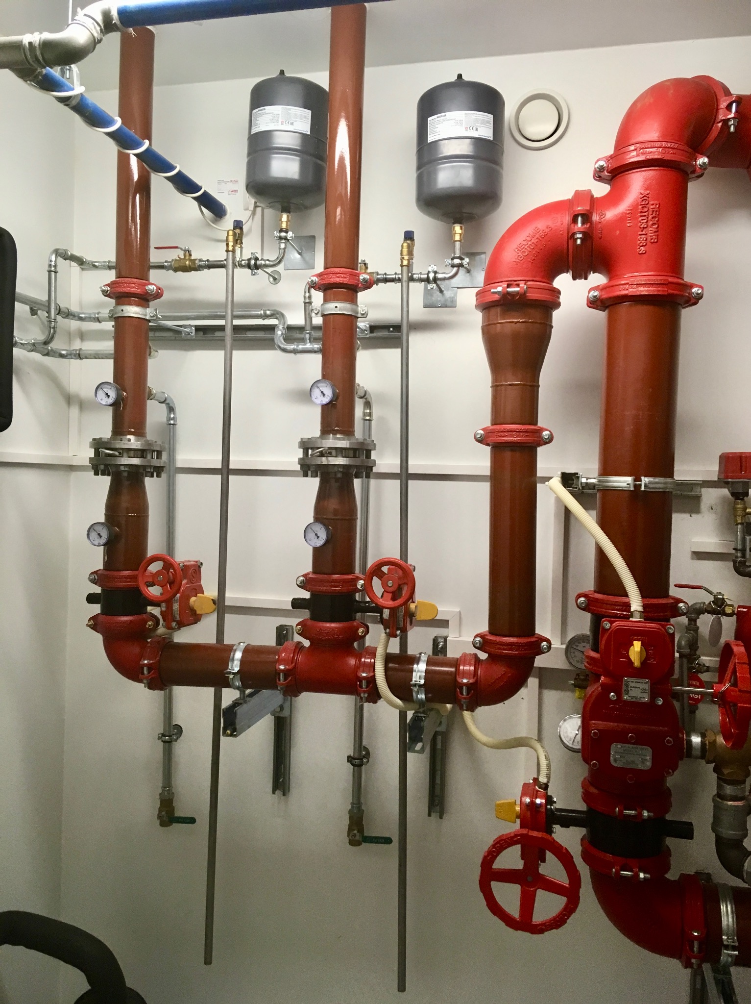 Kompleksowy system rur i zaworów w kolorze czerwonym i brązowym, z manometrami i zbiornikami wyrównawczymi, zamontowany na białej ścianie w pomieszczeniu technicznym.
