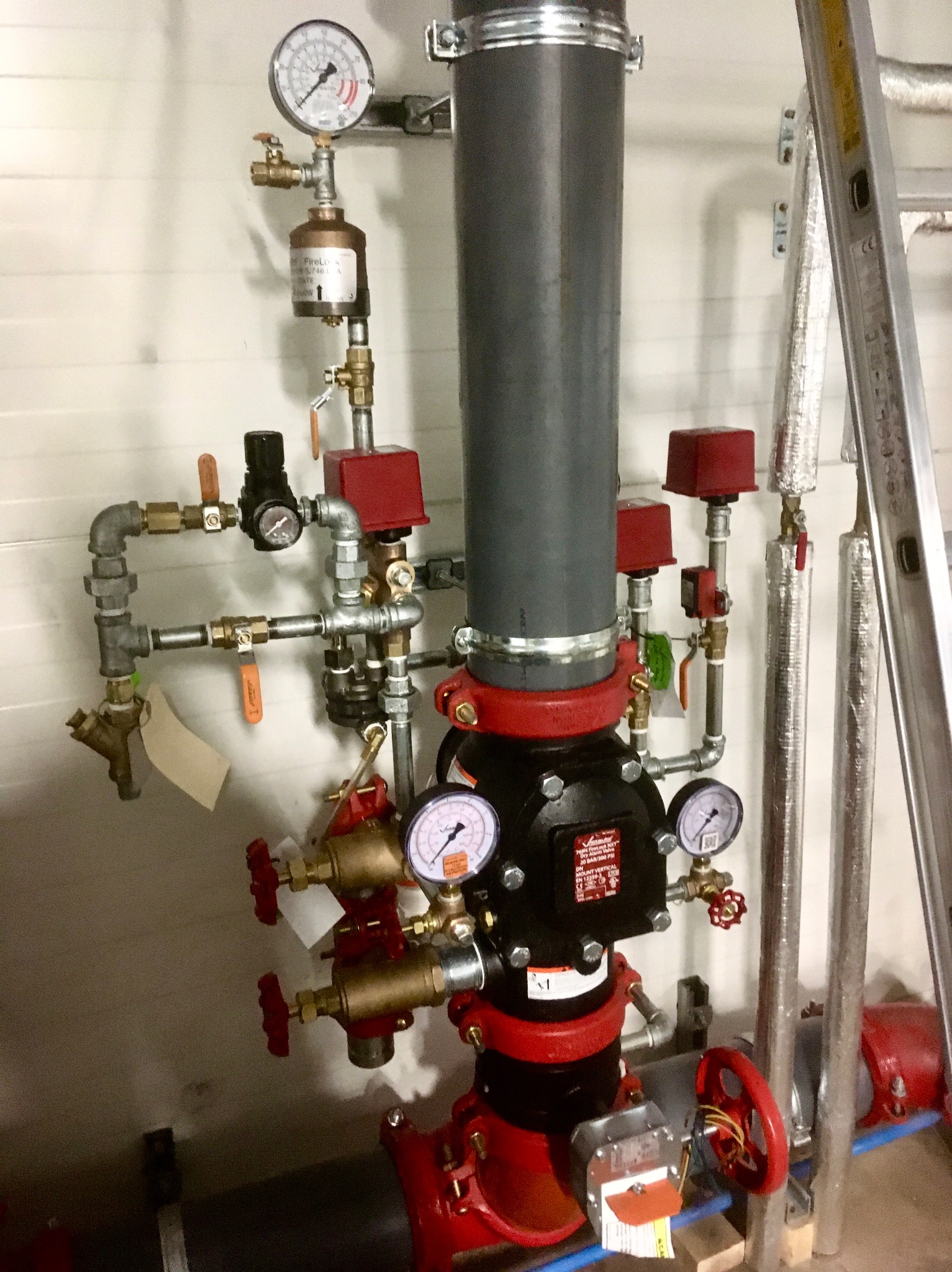 Złożony system rur i zaworów z manometrami w kolorach szarym, czarnym i czerwonym, z widoczną drabinką aluminiową po prawej stronie, prawdopodobnie element instalacji przeciwpożarowej.