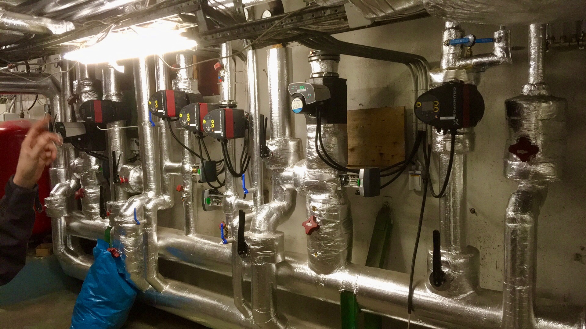 Izolowane rury grzewcze z pompami obiegowymi Grundfos i zaworami, widoczne elementy instalacji w piwnicy, w tle czerwony zbiornik wyrównawczy, instalacja po montażu.