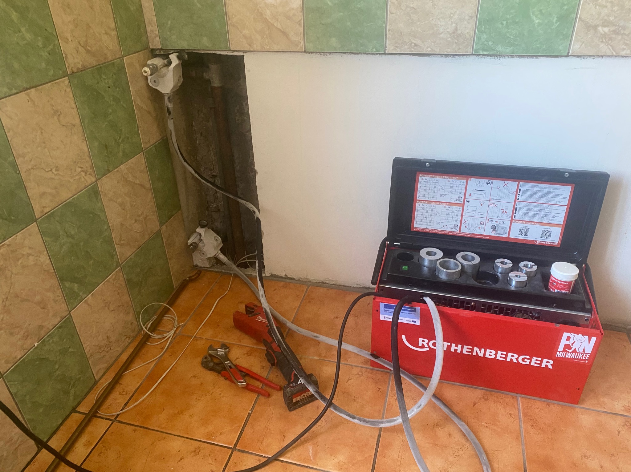 Przyłącze rur w łazience z użyciem zaciskarki Rothenberger i narzędzi Milwaukee, widoczne rury miedziane i plastikowe, płytki ceramiczne na ścianie i podłodze.