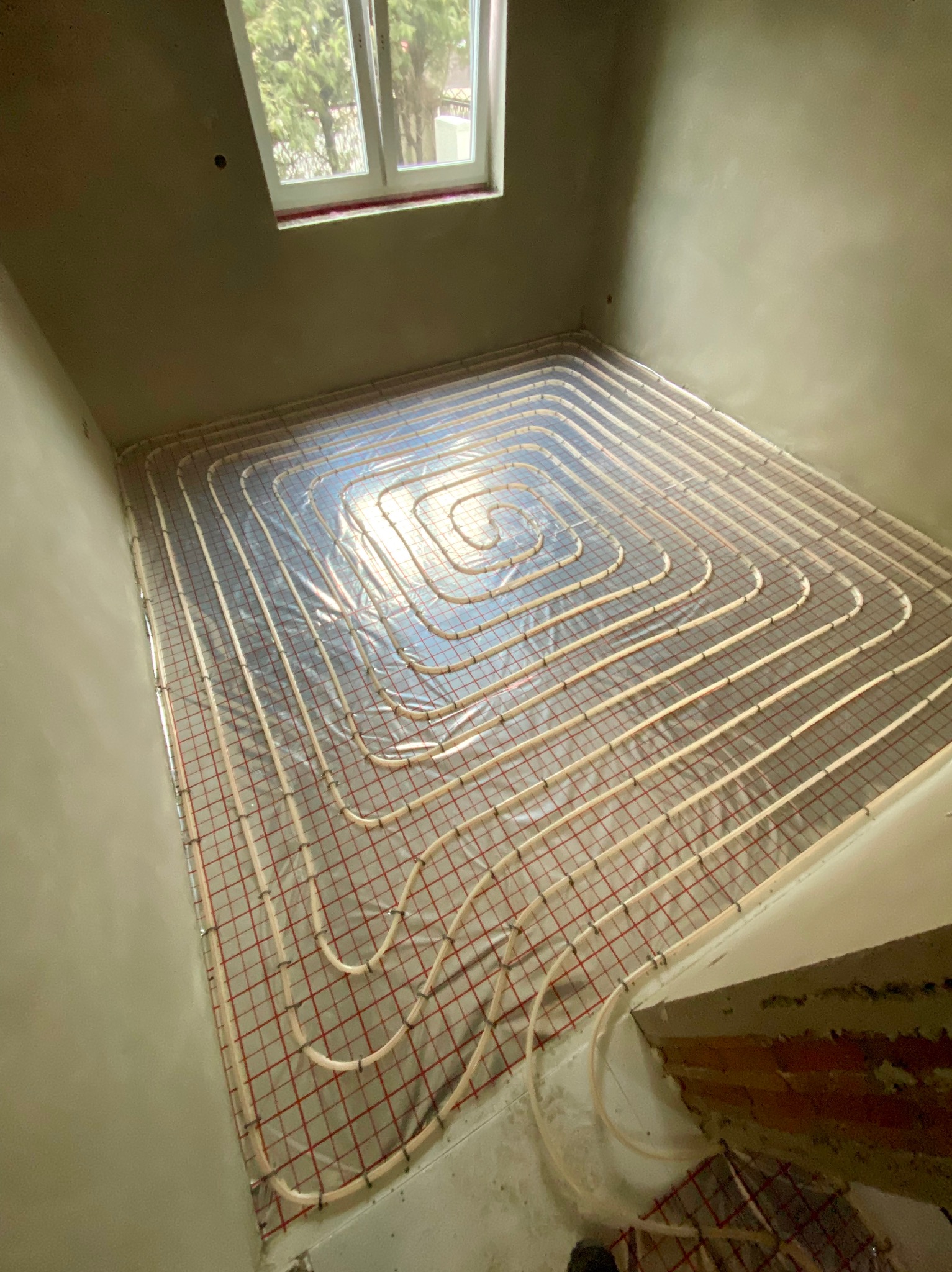 Rury ogrzewania podłogowego ułożone spiralnie na folii izolacyjnej w surowym pomieszczeniu, widok z góry, częściowo otynkowane ściany.