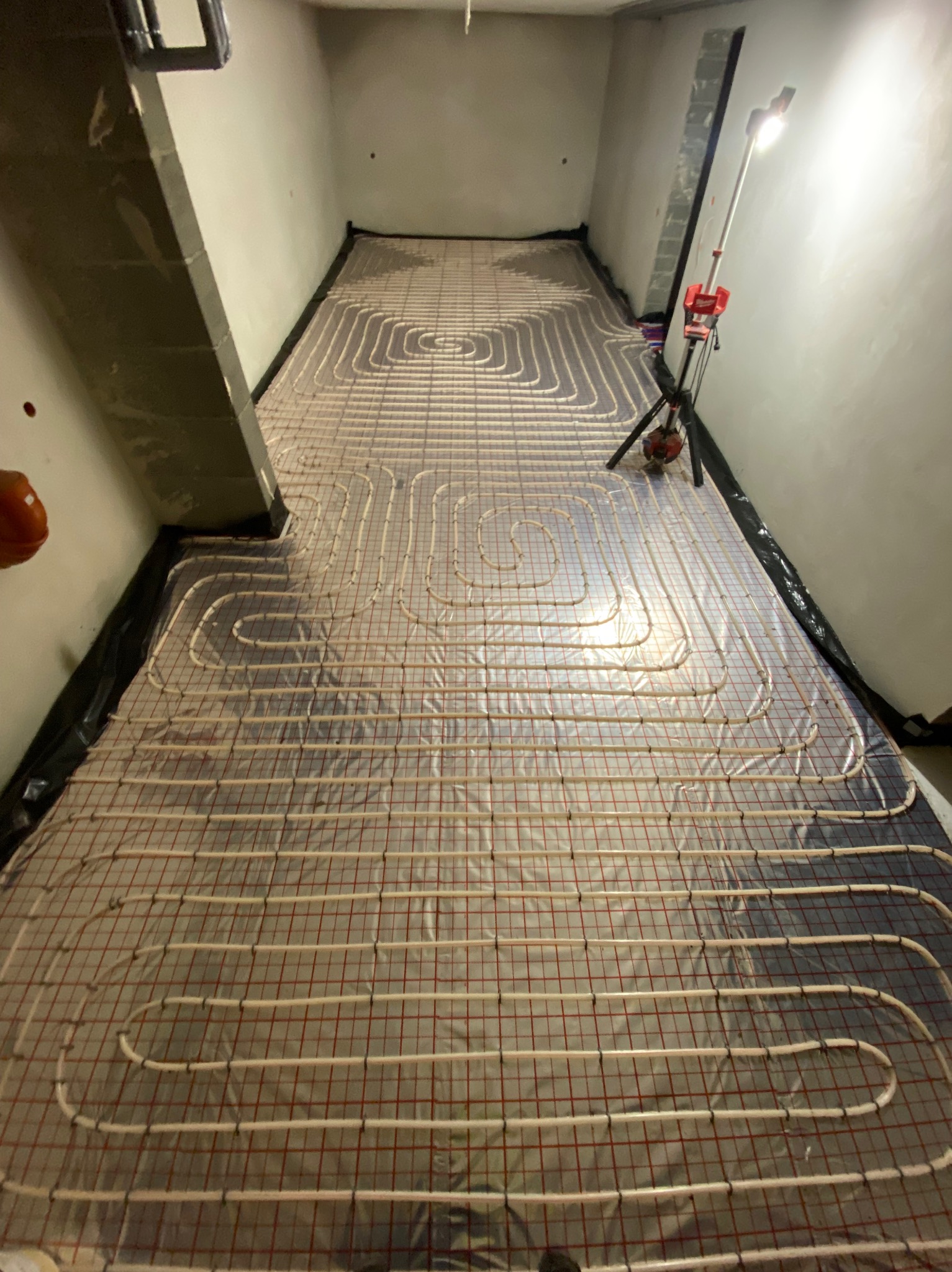 Rozłożona instalacja ogrzewania podłogowego na folii z siatką, widoczna rura grzewcza w kształcie spirali, lampa budowlana oświetla przestrzeń, surowy stan wykończenia.