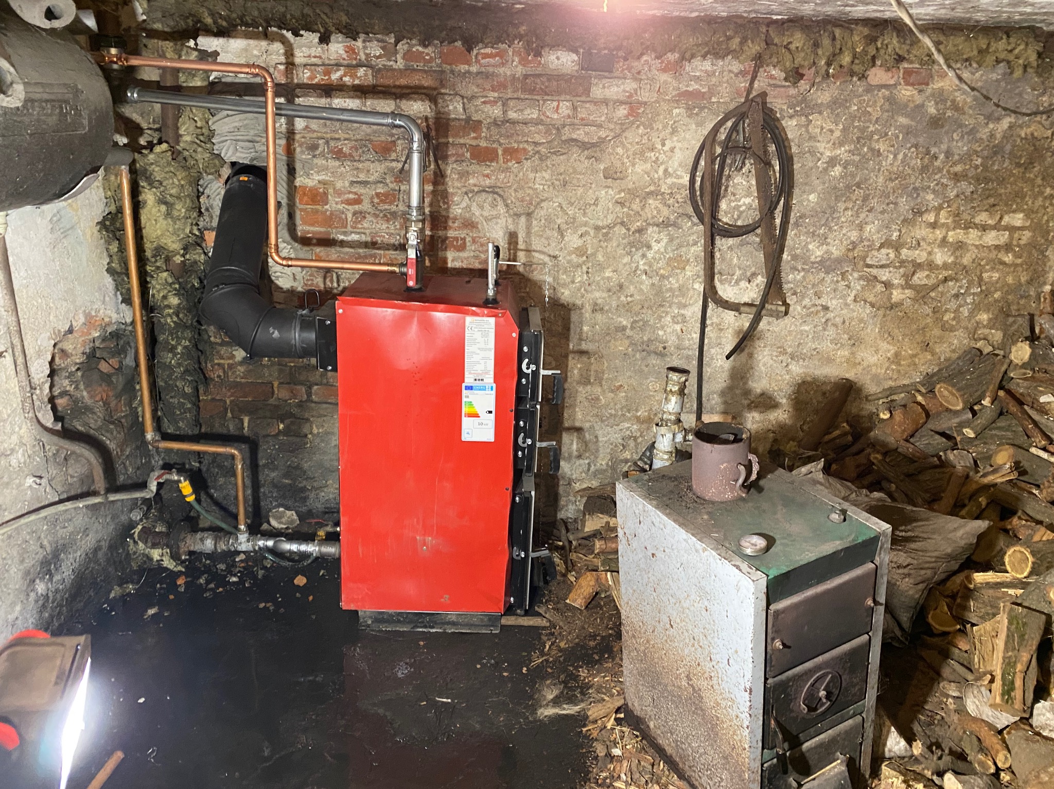 Czerwony piec CO podłączony do instalacji w piwnicy o nierównych ścianach z cegły, w tle stos drewna opałowego i stary piec.