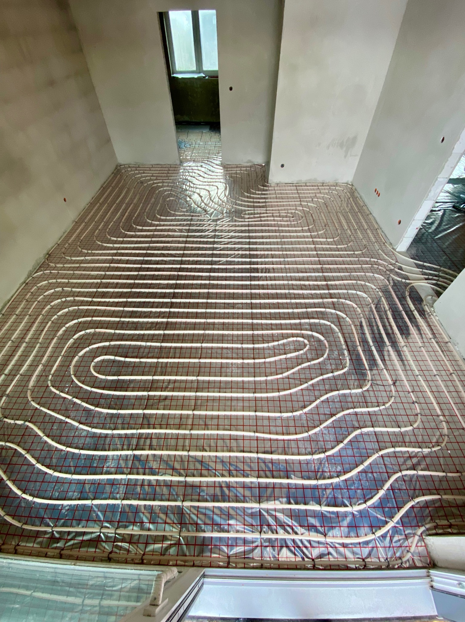 Rozłożona instalacja ogrzewania podłogowego na folii aluminiowej w surowym stanie, widoczne rury w kształcie meandrów i czerwona siatka mocująca, przygotowane do zalania wylewką.