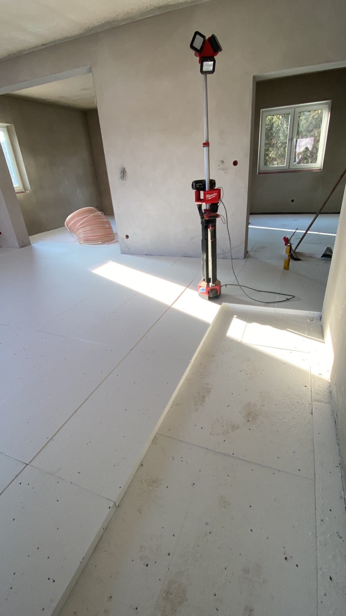 Białe płyty izolacyjne pod ogrzewanie podłogowe ułożone w surowym wnętrzu, widoczna lampa budowlana Milwaukee i zwój rur, ściany w stanie deweloperskim.