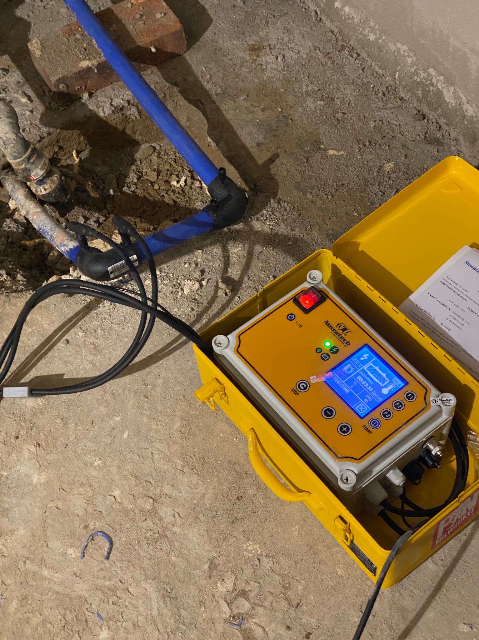 Urządzenie diagnostyczne Nowatech w żółtej skrzynce, podłączone do rur instalacji hydraulicznej w trakcie pomiaru, widoczne parametry na wyświetlaczu.