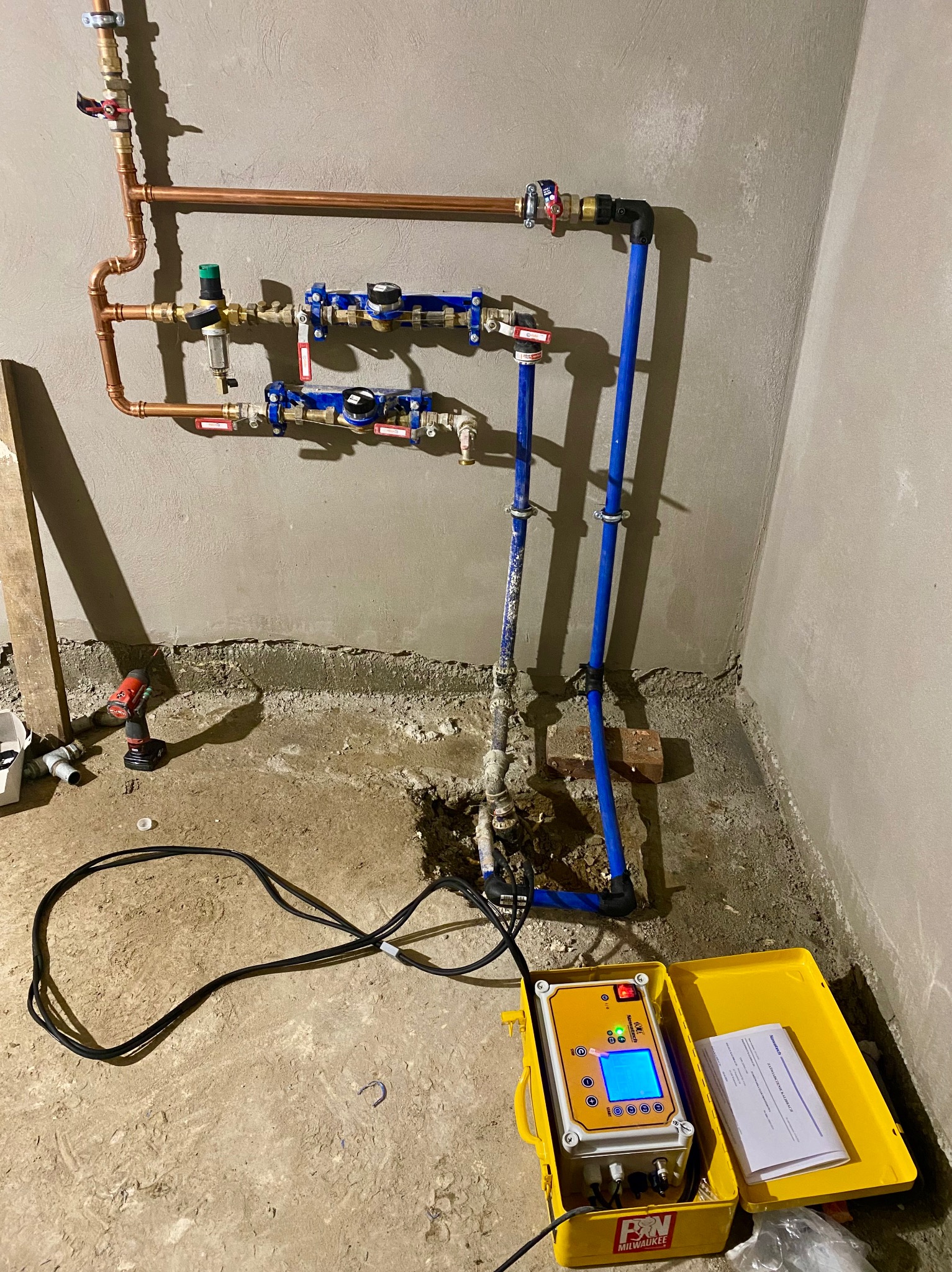 Miedziane i niebieskie rury instalacji wodnej w trakcie montażu, żółta skrzynka z urządzeniem pomiarowym na podłodze, ściana w surowym stanie.