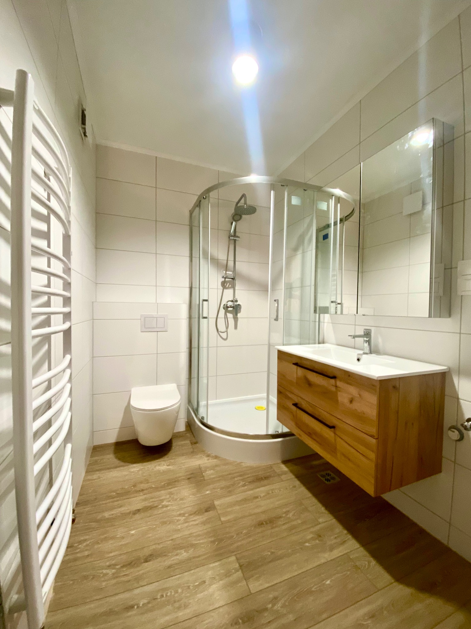 Nowoczesna łazienka z kabiną prysznicową, drewnianą szafką pod umywalką i grzejnikiem drabinkowym, wykończona białymi płytkami i panelami podłogowymi imitującymi drewno.