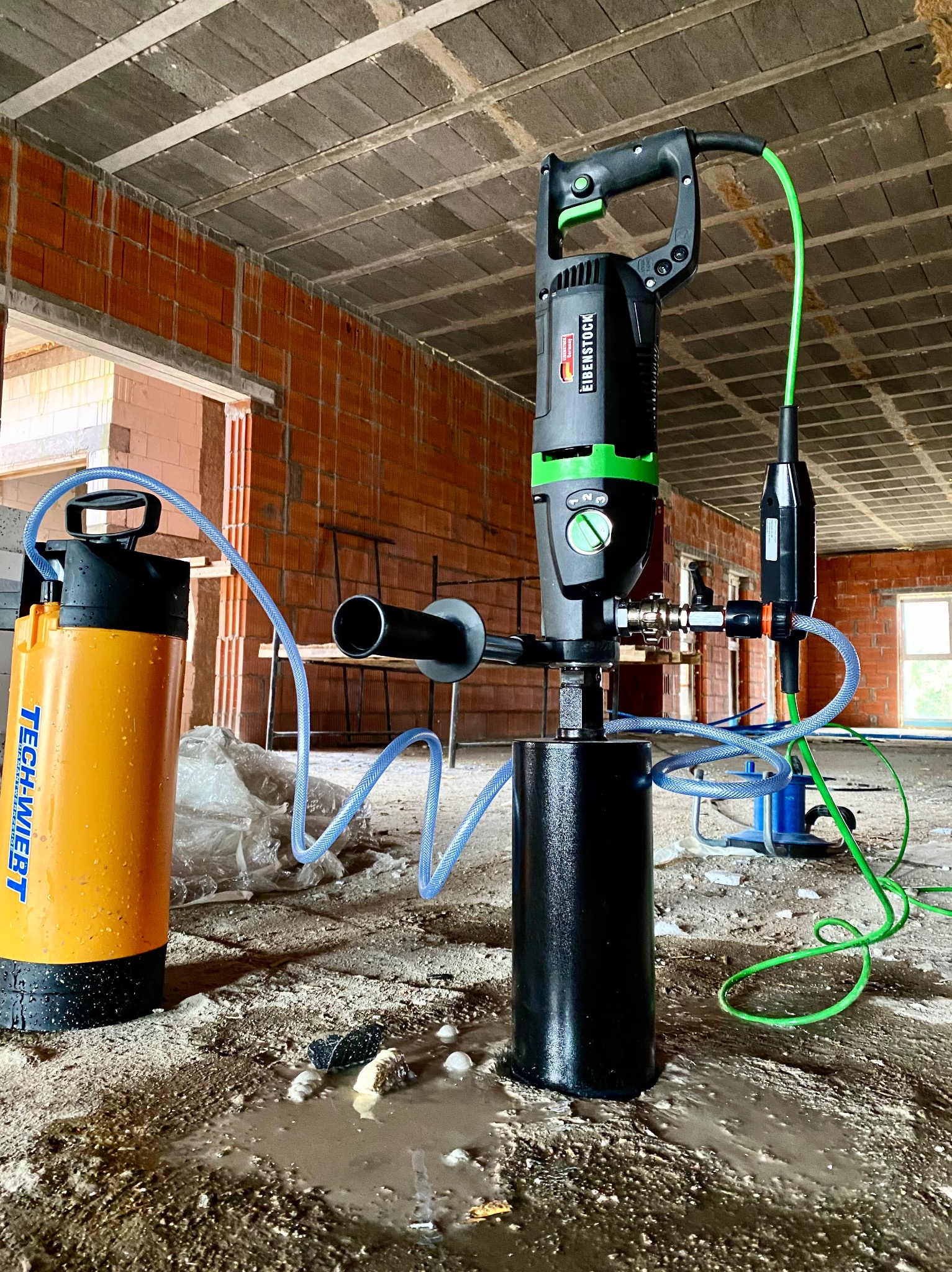 Wiertnica Eibenstock z systemem chłodzenia wodnego podczas wiercenia w posadzce na budowie, widoczne ceglane ściany w tle.