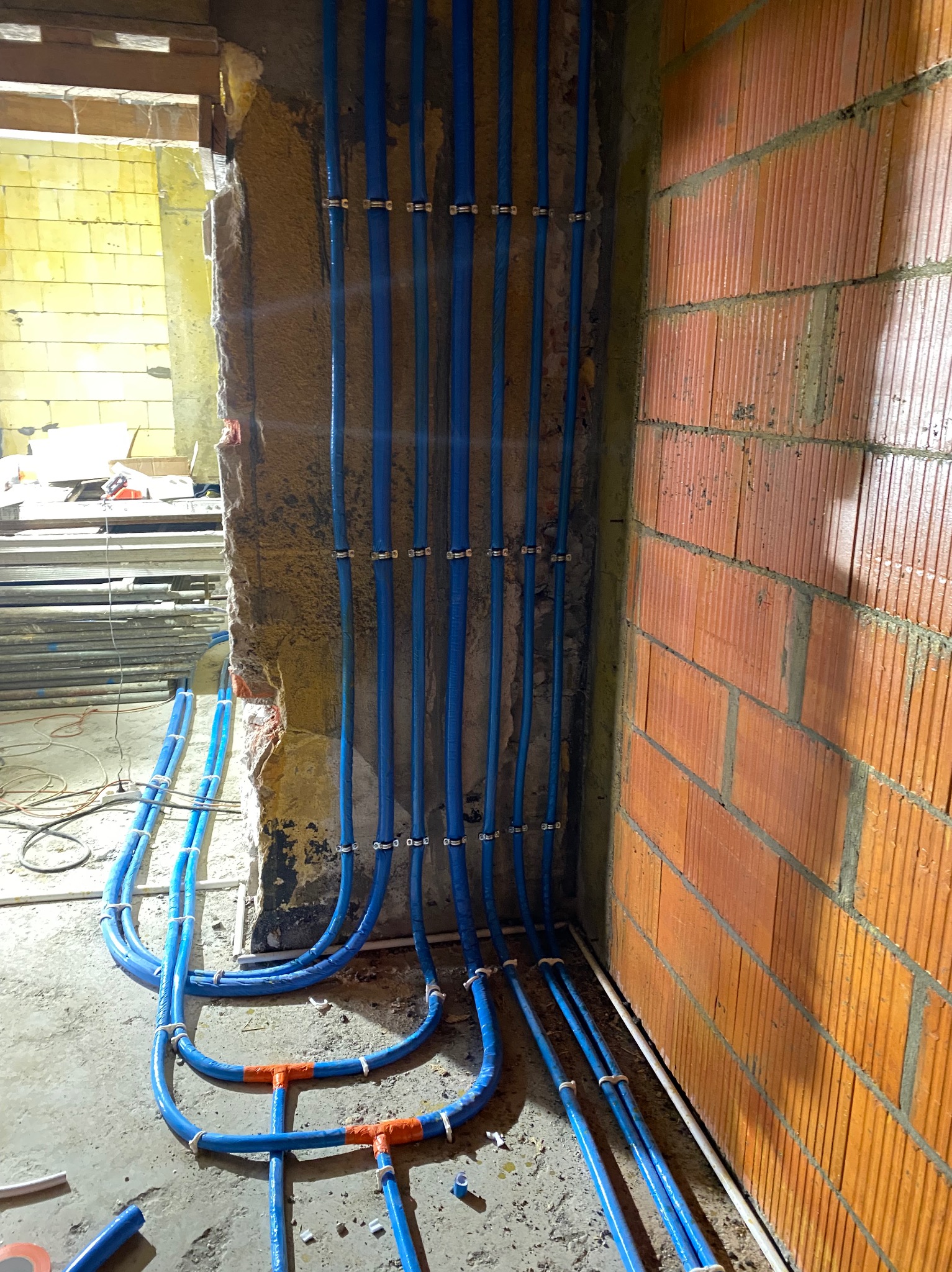 Niebieskie rury hydrauliczne PEX prowadzone wzdłuż surowej ściany z cegły i betonowej ściany z otwartym przejściem, zamocowane metalowymi uchwytami, z widocznymi pomarańczowymi złączkami...