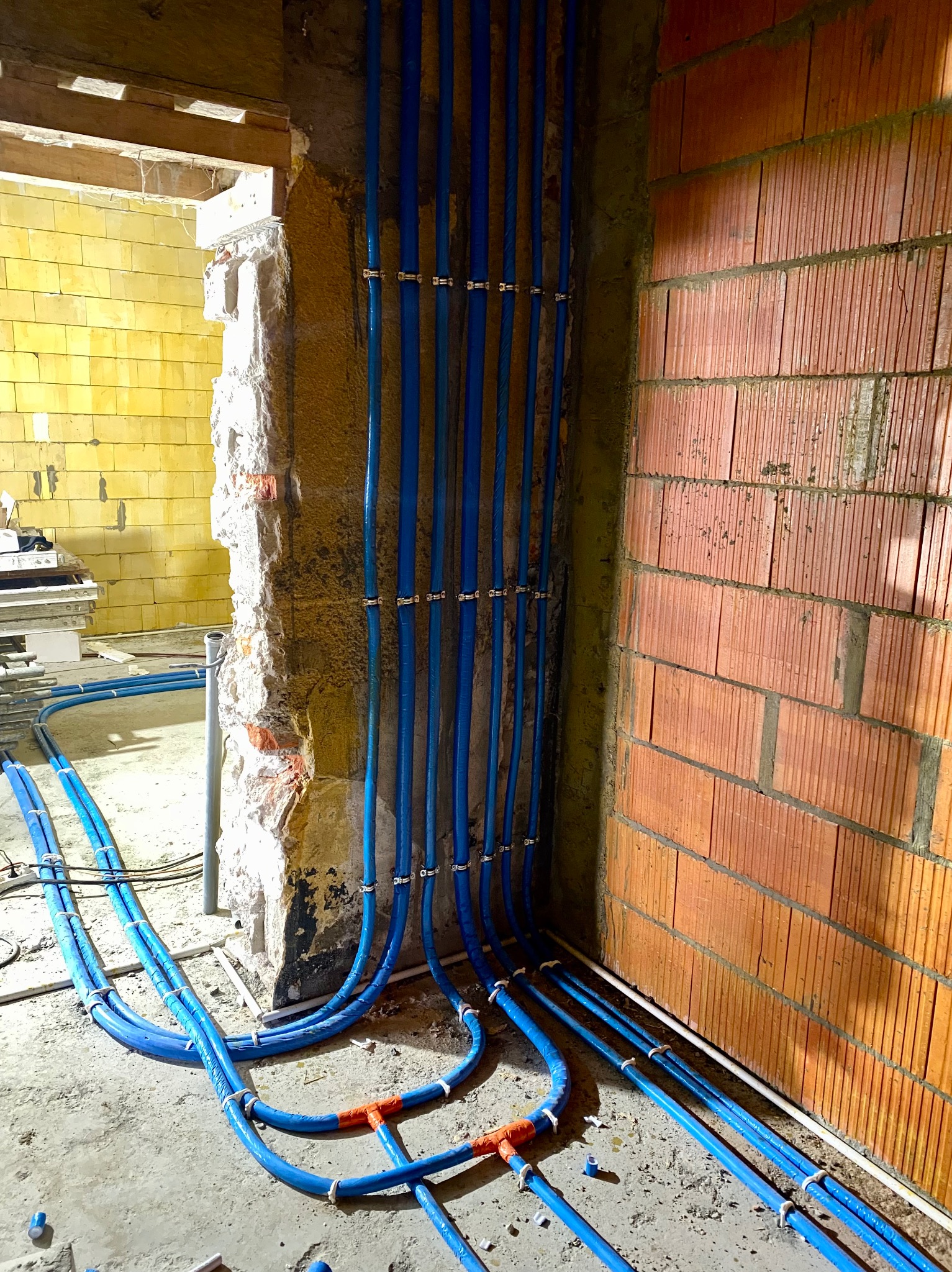 Niebieskie rury instalacji wodnej lub grzewczej, zamocowane do ściany z cegły i surowej ściany z resztkami tynku, widoczne pomarańczowe złączki i metalowe obejmy mocujące.