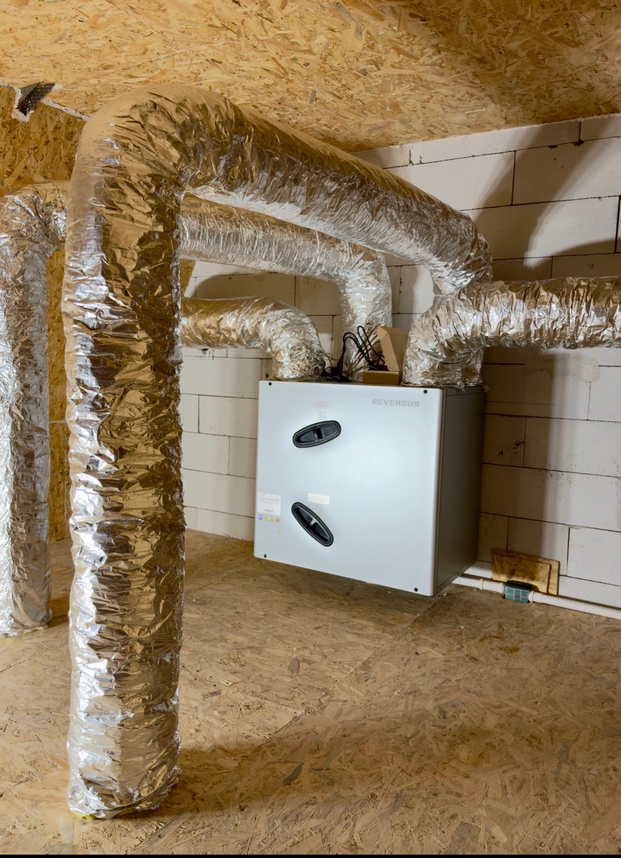 Centrala rekuperacyjna Reversus z aluminiowymi przewodami wentylacyjnymi w surowym wnętrzu. Widoczne białe ściany z bloczków i drewniana podłoga.