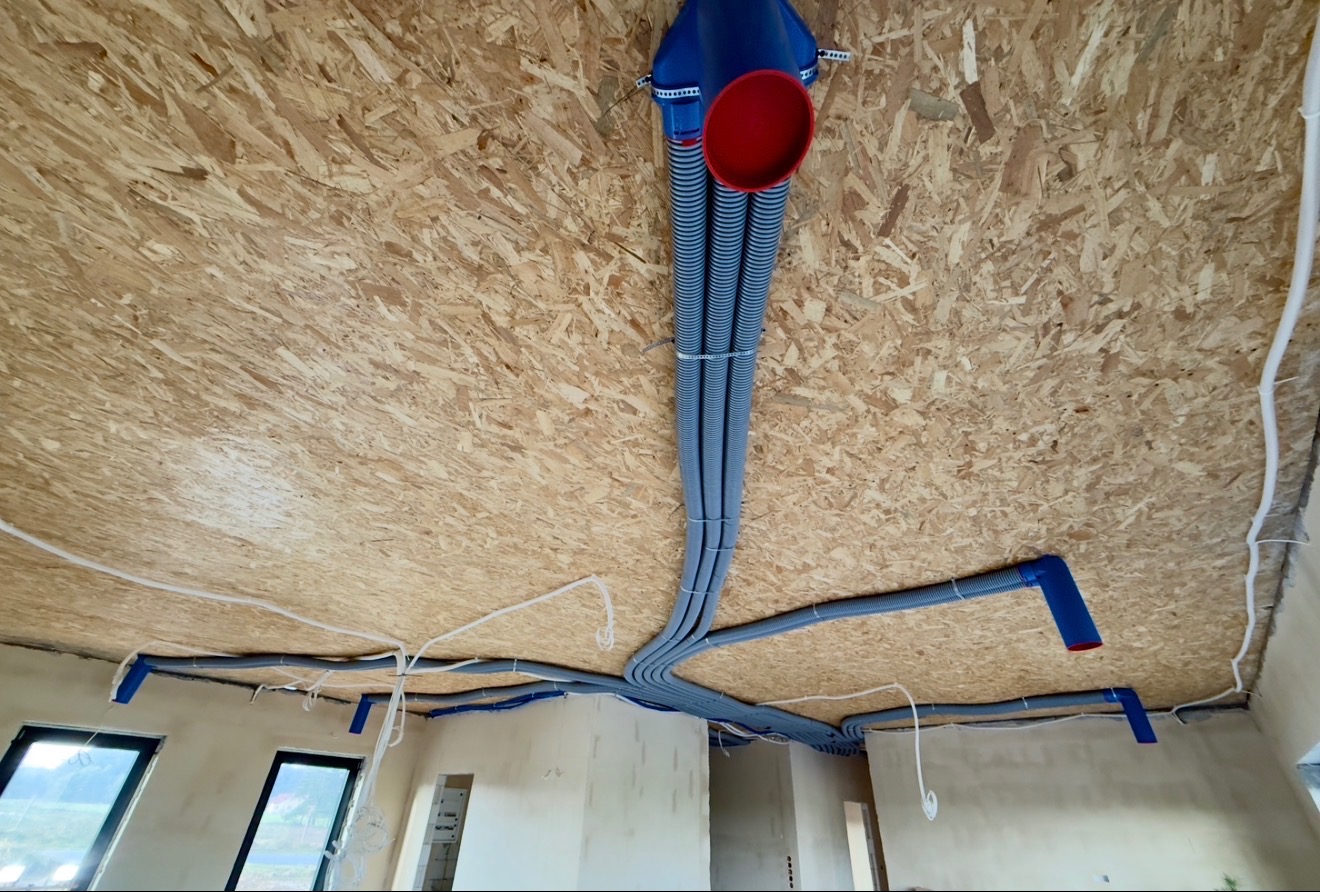 Szare rury wentylacyjne przymocowane do sufitu z płyty OSB w surowym wnętrzu. Widoczne okna i fragmenty ścian. Instalacja w trakcie montażu.
