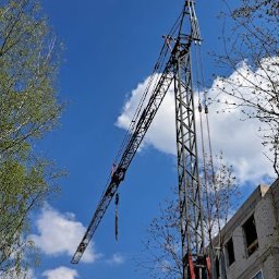 Szkolenia z Pasją Bogumiła Hanert - Żuraw wieżowy w trakcie pracy na budowie budynku mieszkalnego, widoczny na tle błękitnego nieba z białymi obłokami, ujęcie z dołu, częściowo zasłonięty przez gałęzie drzew