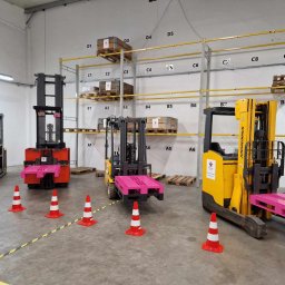 Szkolenia z Pasją Bogumiła Hanert - Wnętrze hali z trzema wózkami widłowymi i pachołkami wyznaczającymi tor przeszkód; na wózkach widoczna różowa paleta; w tle regały z numerowanymi miejscami paletowymi.