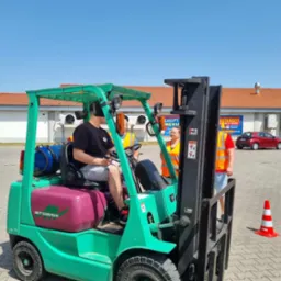 Zielony wózek widłowy Mitsubishi z operatorem w czarnej koszulce, uczestniczącym w szkoleniu, nadzorowany przez instruktorów w pomarańczowych kamizelkach odblaskowych na placu manewrowym.