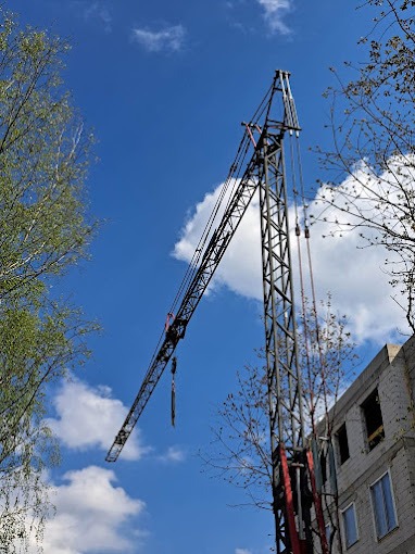 Żuraw wieżowy w trakcie pracy na budowie budynku mieszkalnego, widoczny na tle błękitnego nieba z białymi obłokami, ujęcie z dołu, częściowo zasłonięty przez gałęzie drzew