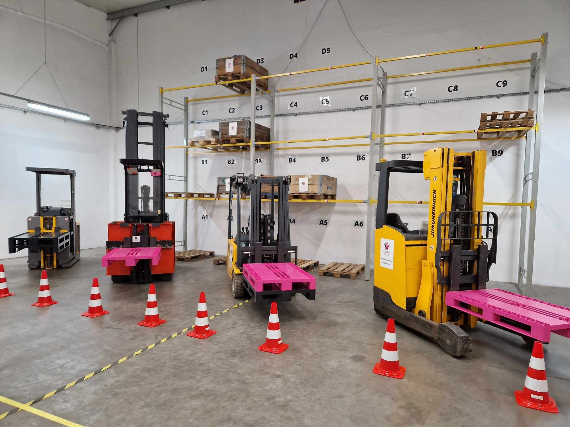 Wnętrze hali z trzema wózkami widłowymi i pachołkami wyznaczającymi tor przeszkód; na wózkach widoczna różowa paleta; w tle regały z numerowanymi miejscami paletowymi.