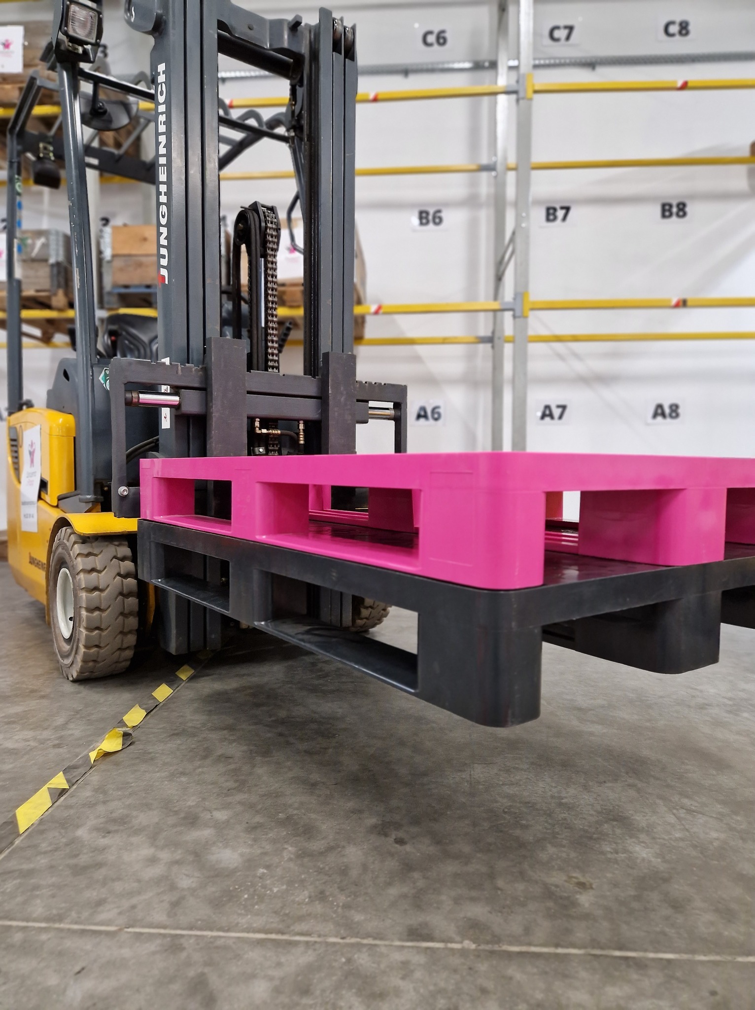 Wózek widłowy Jungheinrich podnosi różową paletę w magazynie z regałami oznaczonymi numerami.
