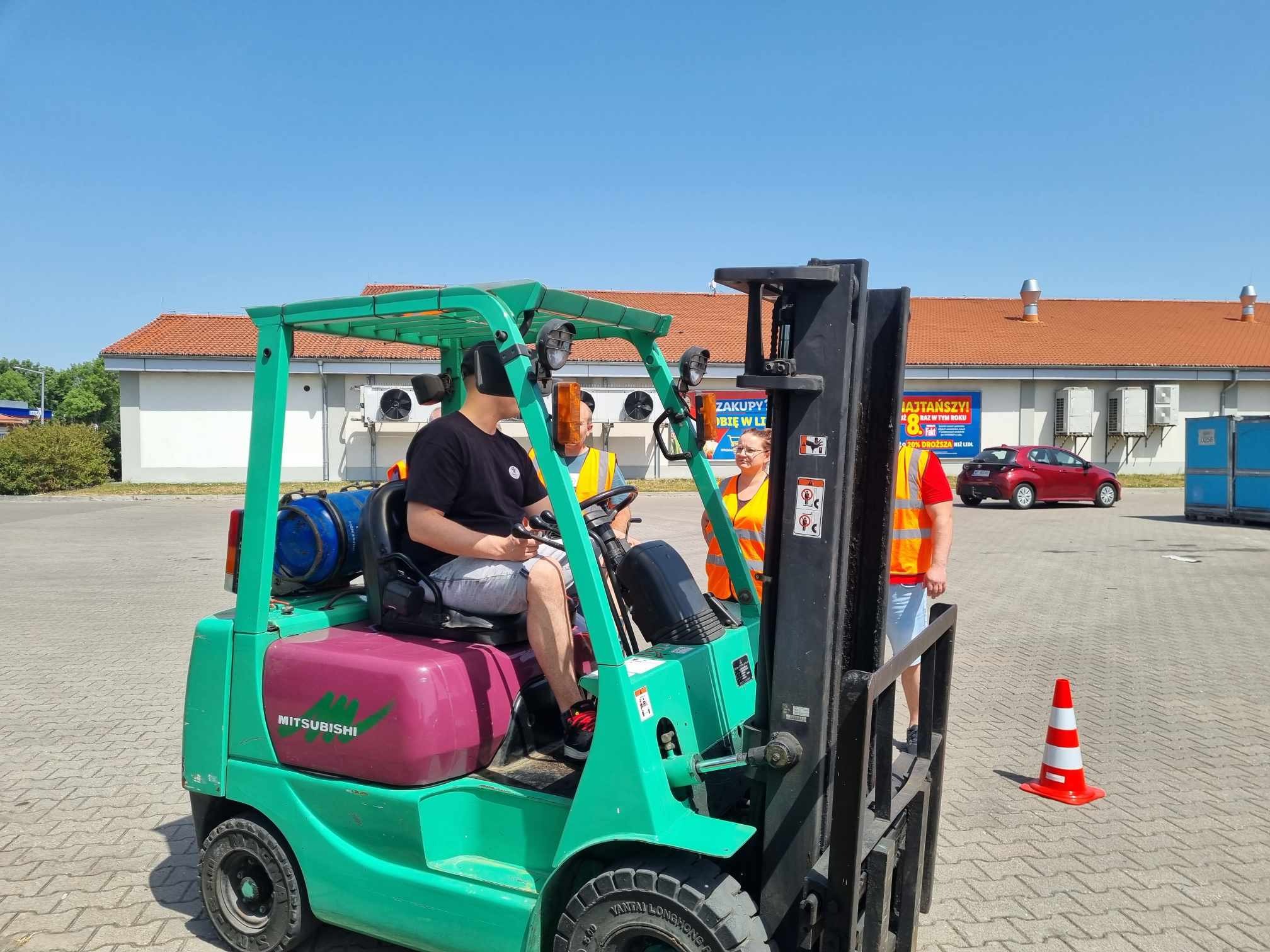 Zielony wózek widłowy Mitsubishi z operatorem w czarnej koszulce, uczestniczącym w szkoleniu, nadzorowany przez instruktorów w pomarańczowych kamizelkach odblaskowych na placu manewrowym.