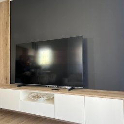 MG Meble na wymiar - Nowoczesna szafka RTV z białymi frontami i drewnianym blatem, na tle ciemnej ściany z drewnianymi lamelami po lewej stronie, telewizor na szafce, robot sprzątający na podłodze.