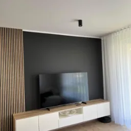 Nowoczesny salon z czarną ścianą, drewnianymi lamelami, białą szafką RTV i telewizorem; widoczny robot sprzątający i fragment fotela.