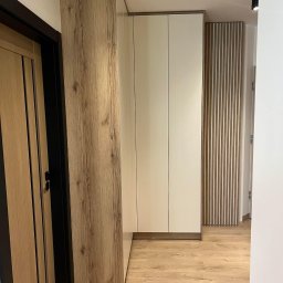 MG Meble na wymiar - Nowoczesna zabudowa przedpokoju z szafą wnękową o białych frontach i drewnianym wykończeniu, dopasowana do koloru drzwi i podłogi, z dekoracyjnym panelem lamelowym w tle.