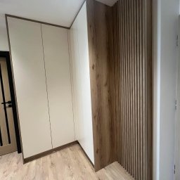 MG Meble na wymiar - Biała szafa wnękowa z drewnianymi akcentami i dekoracyjną ścianką z drewnianych listewek w jasnym wnętrzu.