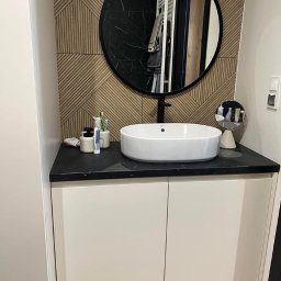 MG Meble na wymiar - Nowoczesna łazienka z okrągłym lustrem, białą umywalką nablatową na czarnym blacie i szafką z białymi frontami. Na blacie kosmetyki i szczoteczki do zębów. W tle ściana z drewnianymi panelami.