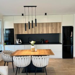 MG Meble na wymiar - Nowoczesna kuchnia z białymi i drewnianymi frontami, czarnym blatem, wiszącymi lampami i stołem z krzesłami o plecionych oparciach.