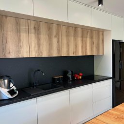 MG Meble na wymiar - Nowoczesna kuchnia z białymi i drewnopodobnymi szafkami, czarnym blatem i backsplash'em, zintegrowanym zlewem i czarną armaturą, oraz Thermomixem po lewej stronie.